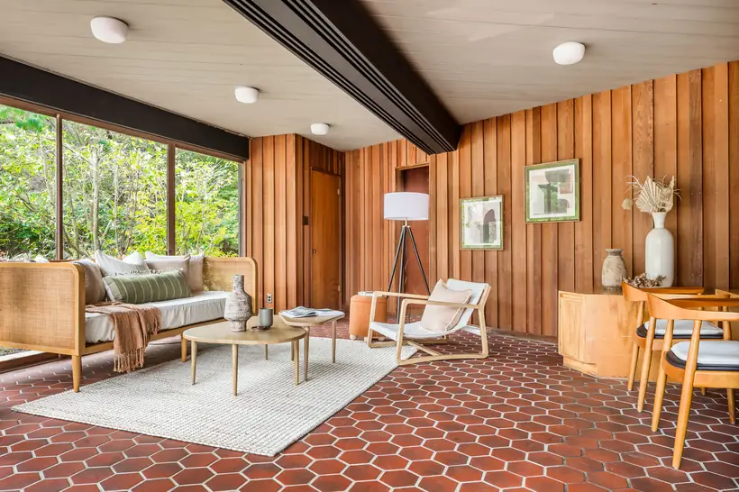 A sitting room floored in terracotta hexagonal tile, walled in vertical redwood, with a wide window onto the trees.